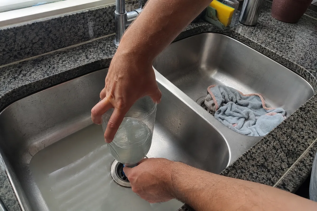 Mãos apertando uma garrafa PET sem fundo encaixada no ralo de uma pia de cozinha de inox, com um pano tampando o ralo da outra cuba